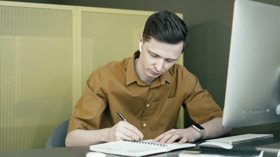 A finance professional taking notes at a desk beside a laptop.